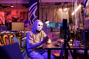 Criminal in anonymous mask recording online fraud video on smartphone. Hacker with hidden identity live streaming threat on mobile phone and asking victim for information on dark web