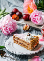 Piece of delicious cake with cream and plum jam on the plate over grey background with fresh plums and peonies flowers. Sweets, dessert and pastry, top view, selective focus