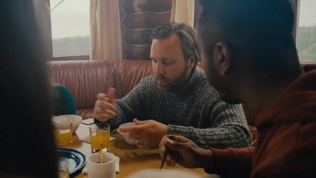 Big multicultural family gather around table, enjoy meal, beverages and discuss their trip. Group of friends resting in holiday home. Travelers relax in wooden cottage during vacation. Tourism.