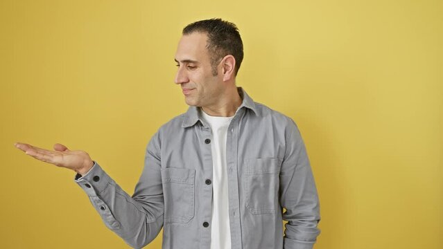 Cheerful, young hispanic guy in shirt, standing and pointing with a welcoming palm gesture. he's happily presenting something, looking directly and smiling for the isolated yellow background shot.