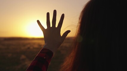 Young girl stretches out her hand to sun, dreams in nature. Sunrise in nature. Hand of happy child girl at Sunset . Prayer in nature. Sunset, sun between fingers of girls hand. Child play hand to sun