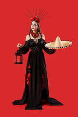 Fototapeta premium Young woman with painted skull on her face, sombrero and lantern against red background. Celebration of Mexico's Day of the Dead (El Dia de Muertos)