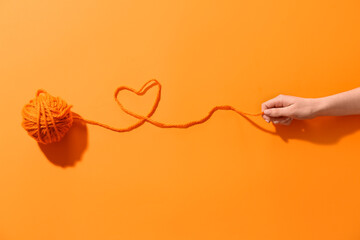 Female hand holding thread of yarn ball with heart on orange background
