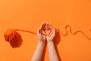 Female hands holding thread of yarn ball with heart on orange background - Powered by Adobe