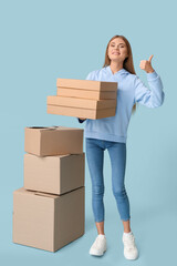 Pretty young woman with cardboard boxes showing thumb-up on blue background. Moving house concept