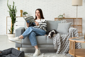 Beautiful young woman with magazine and cute staffordshire terrier puppy sitting on sofa at home - obrazy, fototapety, plakaty