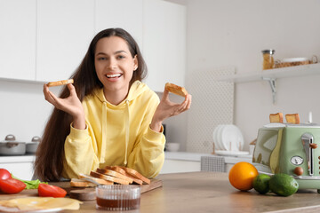 Young beautiful happy woman eating tasty toasts with jam and peanut butter in kitchen