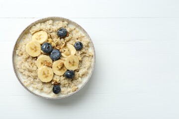 Tasty oatmeal with banana, blueberries, walnuts and honey served in bowl on white wooden table, top view. Space for text