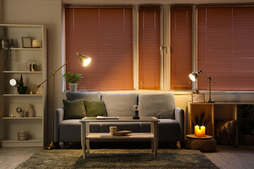 Interior of modern living room with grey sofa, burning candles and glowing lamps at evening