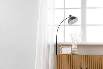 Wooden cabinet with vase, lamp and blank photo frame near window