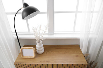 Wooden cabinet with vase, lamp and blank photo frame near window