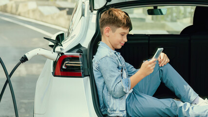 Little boy sitting on car trunk, using smartphone while recharging eco-friendly car from EV...
