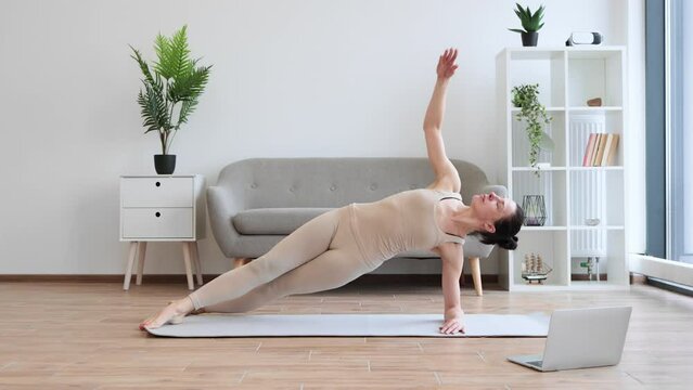 Healthy senior female practising modern yoga with Vasisthasana exercise at home. Beautiful calm yogini in beige activewear improving body strength while holding Side Plank Pose.