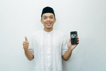 Indonesian Asian muslim man smiling and showing smartphone with thumb up wears black hat called...