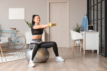 Fototapeta na wymiar Sporty young woman with dumbbell training on fitball at home