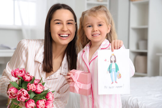 Little girl congratulating her happy mom with bouquet of beautiful flowers and greeting card in bedroom. Mother's Day celebration