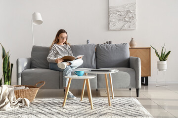 Young woman in eyeglasses reading magazine on grey sofa at home