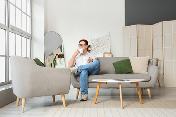 Young man in eyeglasses with cup of tea and mobile phone sitting on grey sofa at home