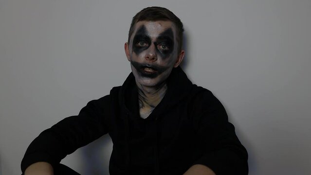 Young Caucasian man with black clown Halloween makeup sitting down and talking to the camera
