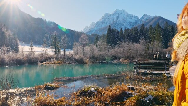 4K video of a young caucasian female standing near the lake in the natural reserve - Zelenci, Slovenia. Observing scenery with yellow winter jacket which adds a perfect contrast to snowy outdoor scene