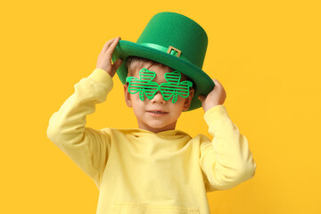 Cute little boy in leprechaun hat and decorative glasses in shape of clover on yellow background. St. Patrick's Day celebration