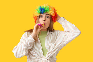 Beautiful young woman in funny clown disguise with party whistle on yellow background. April Fools...