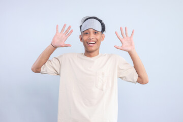 Young Asian guy wear pajamas sleep and eye mask raising hands with surprised expression