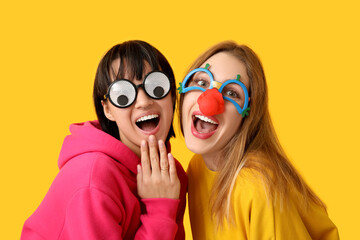 Beautiful young happy women in funny disguise on yellow background. April Fools Day celebration - obrazy, fototapety, plakaty