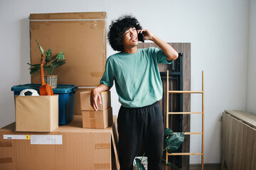 Young Asian curly man standing by boxes and talking on mobile phone at new home. Moving, people and real estate concept