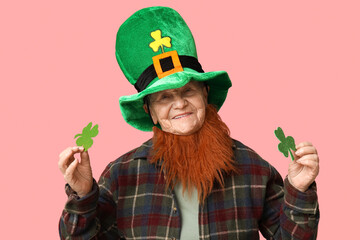 Senior woman in leprechaun hat with red beard holding clover on pink background. St. Patrick's Day...