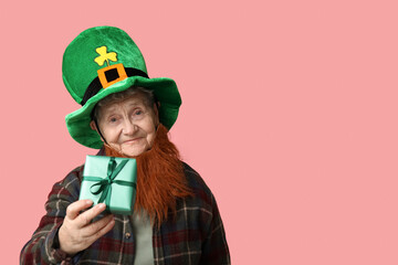 Senior woman in leprechaun hat with red beard holding gift box on pink background. St. Patrick's Day celebration