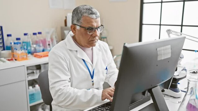 Mature, gray-haired man in lab coat working at computer in a laboratory setting with microscope and test tubes.