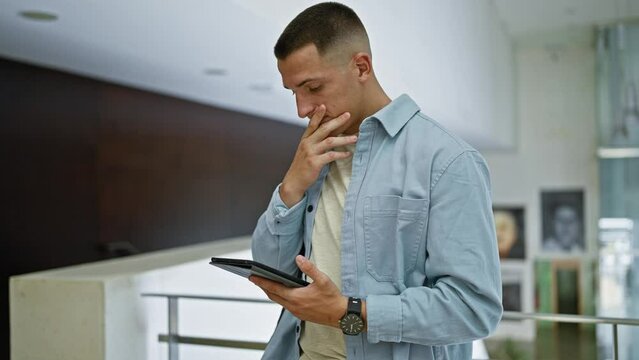 A thoughtful young man indoors, holding a tablet with a concerned expression, wearing a denim shirt and a watch.