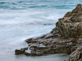 A serene and dynamic scene of a rocky shoreline and the ocean. A series of jagged, brown rocks protrude into the sea, appearing wet and rugged.