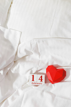 Valentine's Day concept, red paper valentine heart and 14 february holiday date on wooden calendar in bed on on  white crumpled sheets. Minimal style flat lay photo, top view, copy space