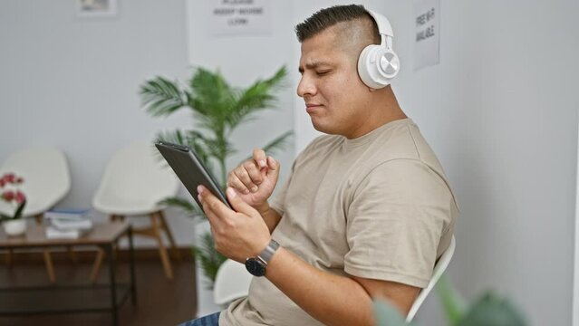 Handsome young latin man chilling out, effortlessly dancing in his chair, while listening to an intoxicating song on his touchpad in a quiet waiting room