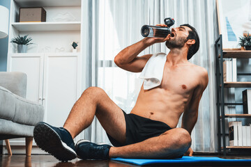 Athletic and sporty man drinking water on fitness mat after finishing home body workout exercise...