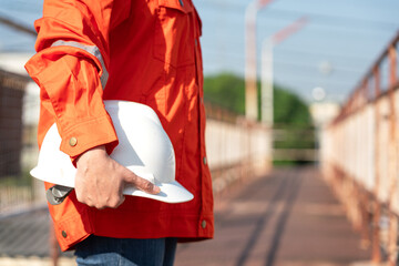 An engineer or construction worker is holding a white safety helmet, wearing orange coverall, standing on the working platform walkway. Ready to work in the challenge workplace concept scene. 