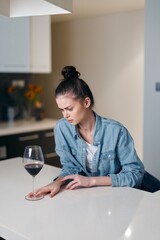 Lonely woman sitting alone at the kitchen table, holding a glass of wine.