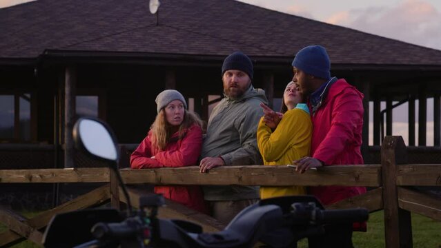 Group of multiethnic tourists stand near wooden fence, talk and look at beautiful sunset. Hiking friends relax during vacation trip in mountains. Stylish wooden cottage in the background. Slow motion.
