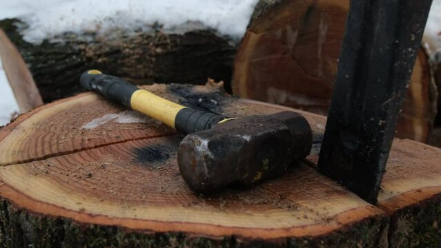 Close up of large hammer and iron wedge prepared to be used to split tree stump while moving camera away exposing more large logs covered in snow.