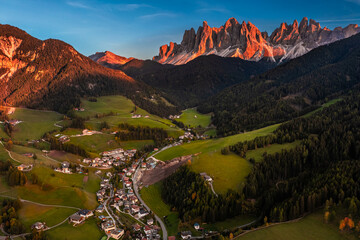 Val Di Funes, Dolomites, Italy - Aerial view of the Val di Funes province at South Tyrol with St. Johann church in San Valentino, the Italian Dolomites in warm sunset colors and blue sky at background