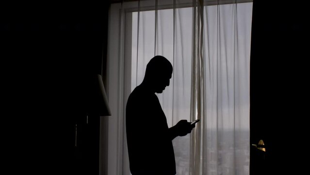 Silhouette of a man standing near the window with the curtains closed, typing on a mobile phone screen, the concept of privacy and anonymity.