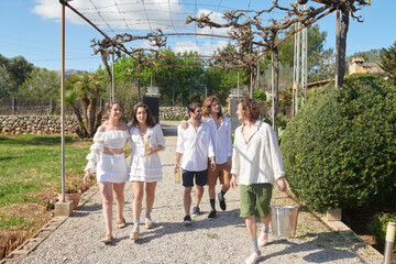 A lively group of friends walks through a country estate, with two women leading the way, one holding a bottle, followed by three men, one carrying a bucket, all in high spirits