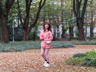 Beautiful young Chinese woman smiling and walking in autumn forest, wearing pink loose oversized top with small white shoulder bag. Emotions, people, beauty, youth and lifestyle portrait.