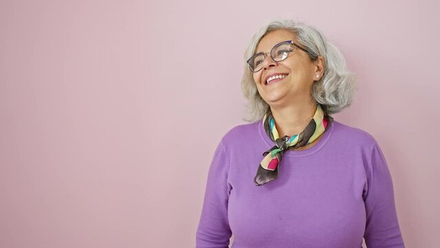 Confident grey-haired woman with glasses stands laughing, a beaming smile on her face as she looks off to the side over a pink isolated background, emulating great natural expression.