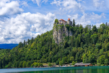 Fototapeta na wymiar Castle on top of rock with waving Slovenian flag at lakeshore of Slovenian Lake Bled on a blue cloudy summer day. Photo taken August 8th, 2023, Bled, Slovenia.