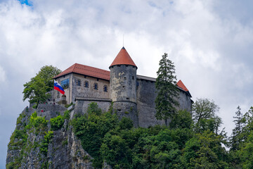 Fototapeta na wymiar Scenic view of castle on top of rock with Slovenian flag waving at lakeshore of Slovenian Lake Bled on a cloudy summer day. Photo taken August 8th, 2023, Bled, Slovenia.