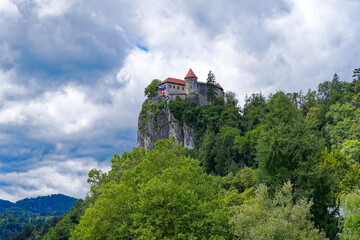 Fototapeta na wymiar Scenic view of castle on top of rock with Slovenian flag waving at lakeshore of Slovenian Lake Bled on a cloudy summer day. Photo taken August 8th, 2023, Bled, Slovenia.