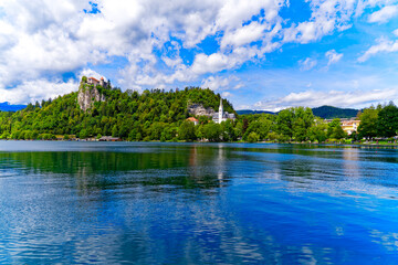 Obraz premium Scenic view of Lake Bled with beautiful reflections and castle on a rock and woodland in the background on a blue cloudy summer day. Photo taken August 8th, 2023, Bled, Slovenia.